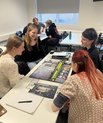 Four students sit around a table in a classroom, smiling and discussing while playing a board game with cards and tokens; other students work at tables in the background.