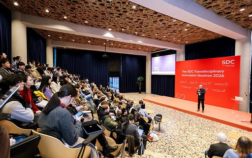 Wide shot of the auditorium at the SDC Transdisciplinary Innovation Ideathon 2026 in Beijing. Students and staff are seated in tiered rows facing a stage with a large red event backdrop and a speaker presenting.